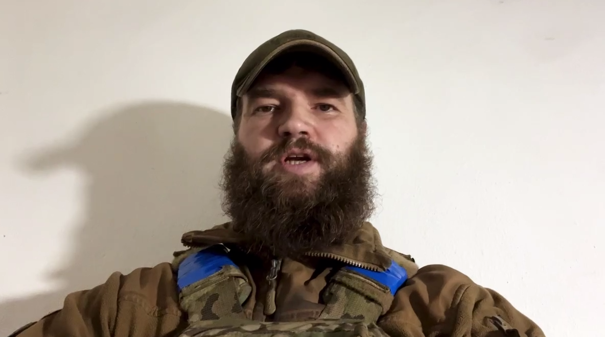 A soldier in a cap and body armour recording a video address.
