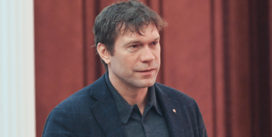 A man in a dark jacket in front of red curtains.